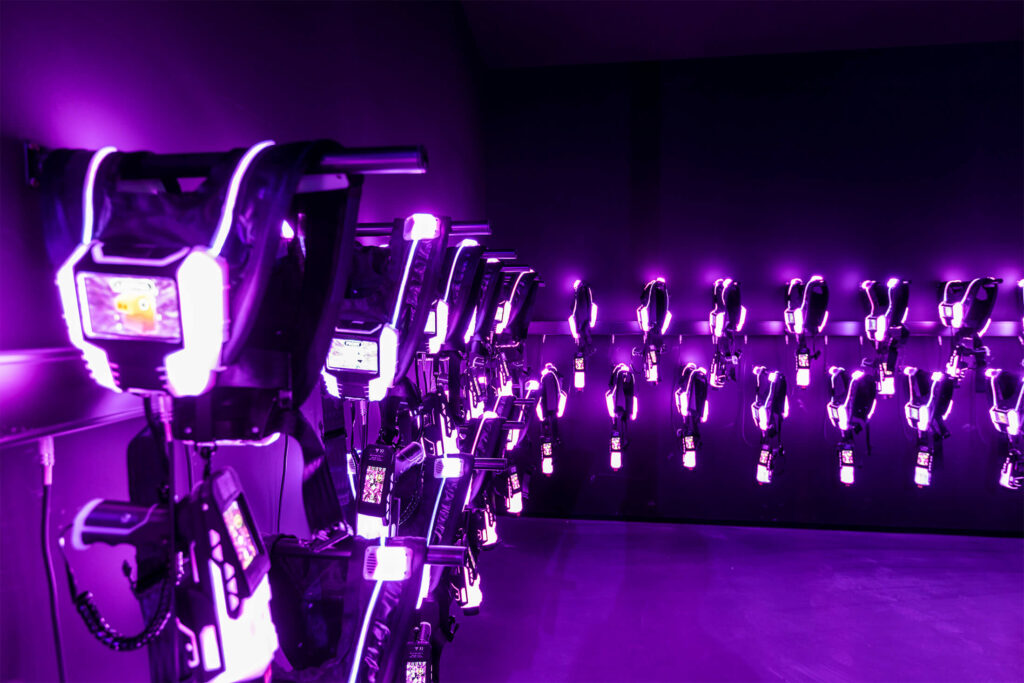 Eine Menge Lasertag-Westen und die zugehörigen Waffen leuchten violett an der Wand.